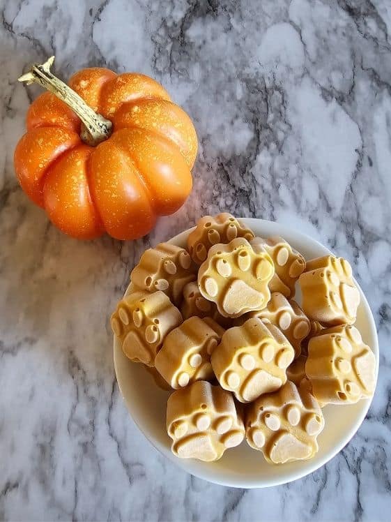 Paw shaped peanut butter pumpkin dog treats in a white bowl next to a small pumpkin.