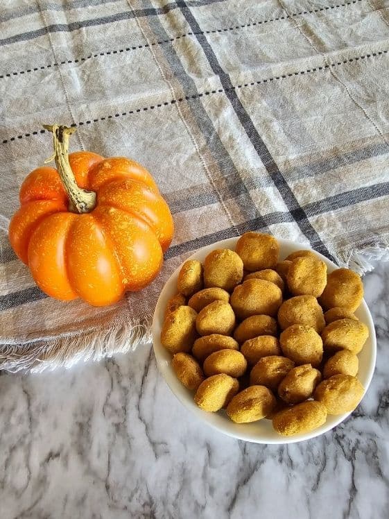 Baked dog treats in a white bowl next to a small pumpkin.