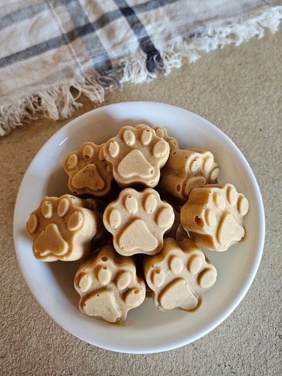 A bowl of frozen dog treats shaped like paw prints, made with banana and peanut butter, sits on a countertop alongside a textured cloth.