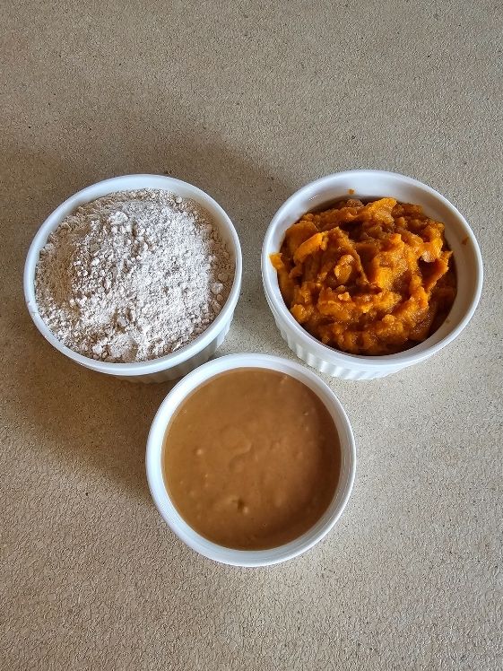 Whole wheat flour, pumpkin puree, and peanut butter in individual white bowls.