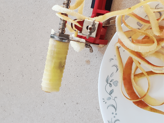 The core of an apple on the apple peeler, corer, slicer with peel laying on a plate next to it.