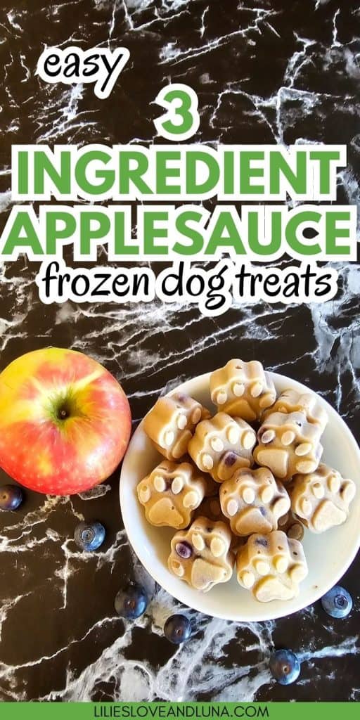 A bowl of homemade frozen dog treats shaped like paw prints sits beside an apple and some blueberries on a marbled surface, with text indicating they are easy three-ingredient applesauce treats.