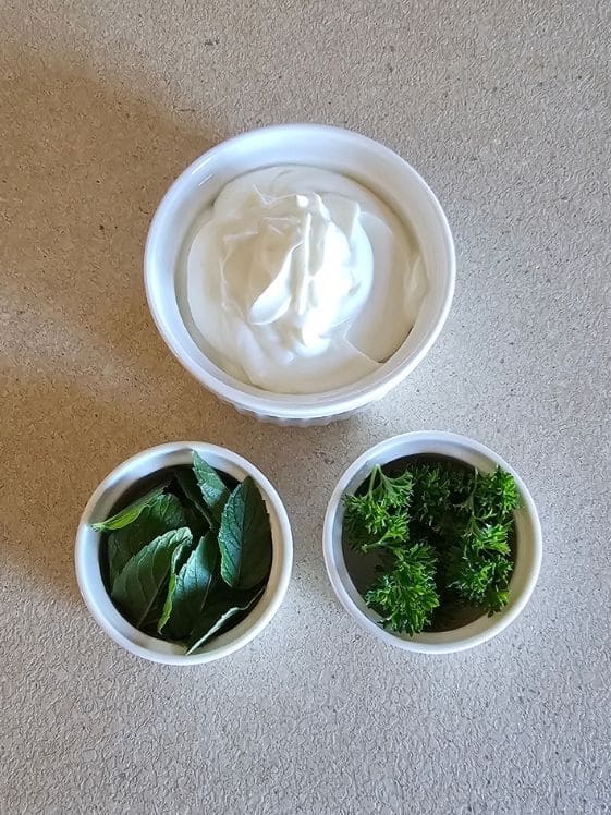 Plain Greek yogurt, mint leaves, and parsley leaves in individual white bowls.
