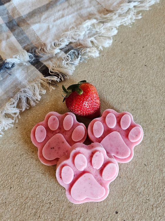 Paw shaped strawberry ice cream for dogs next to a fresh strawberry and linen towel.