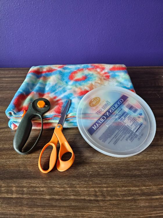 A fleece blanket, a rotary cutter, scissors, and ice cream pail lid laying on a table.
