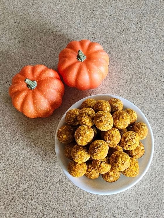 Two small pumpkins next to a white bowl filled with no bake peanut butter pumpkin dog treat balls.