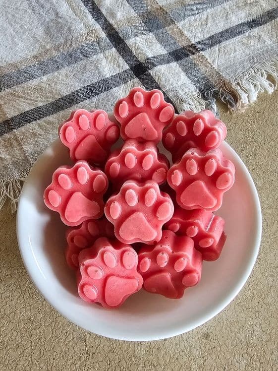 Paw shaped strawberry pineapple frozen dog treats in a bowl next to a linen towel.