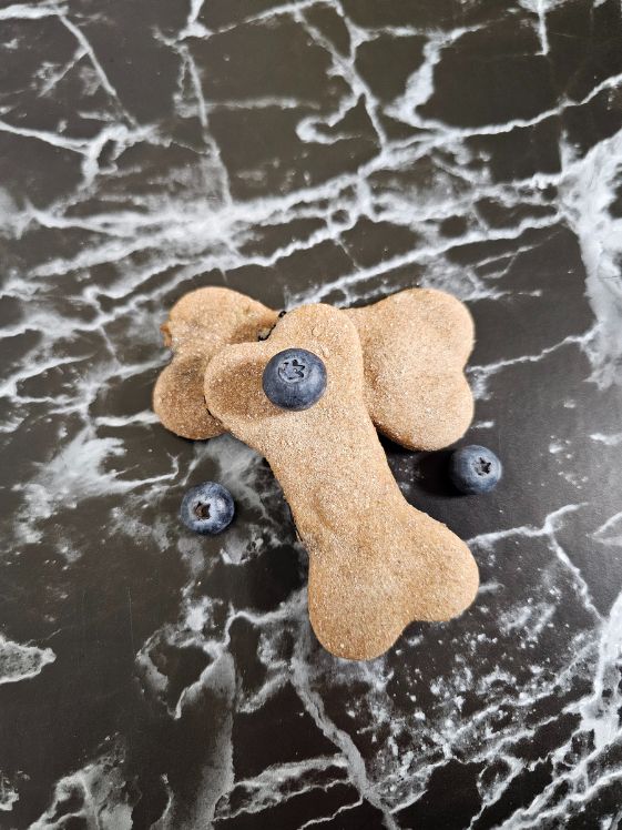 Two bone shaped blueberry dog treats on a black marble background with blueberries around them.