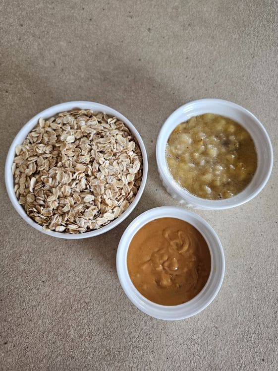 A bowl of old fashioned oats, a bowl of mashed banana, and a bowl of peanut butter on a counter top.