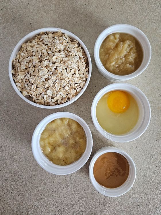 Oats, mashed banana, an egg, applesauce, and peanut butter in individual white bowls.