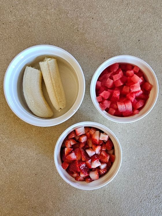 Banana, watermelon, and strawberries in individual white bowls on the counter.