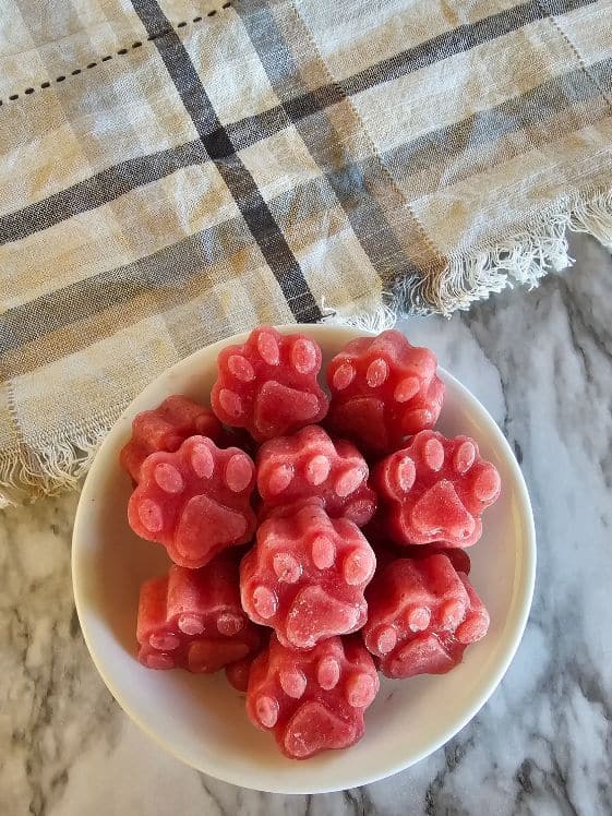 A bowl of paw shaped banana melon berry frozen dog treats next to a kitchen towel.
