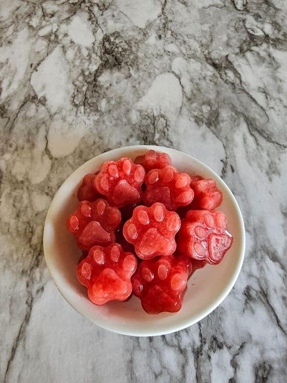 Paw shaped frozen strawberry watermelon dog treats in a bowl on a marble surface.