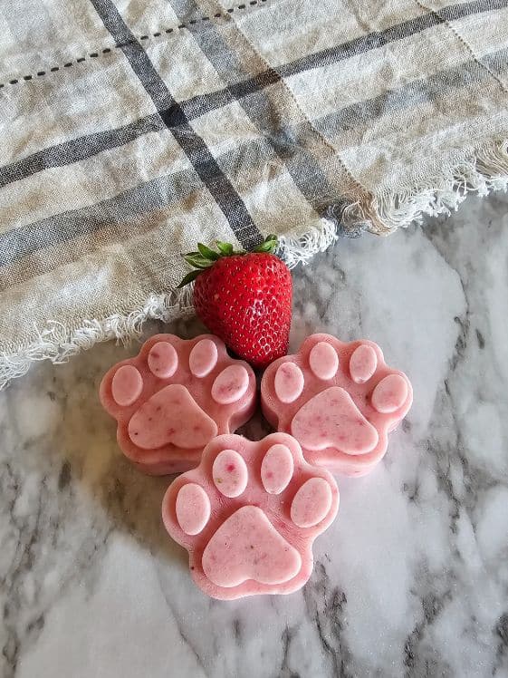 Three paw shaped strawberry watermelon ice cream dog treats next to a strawberry and a kitchen towel.
