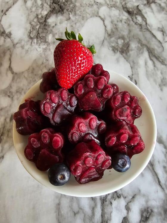 Paw shaped watermelon berry frozen dog treats in a bowl with a strawberry and some blueberries.