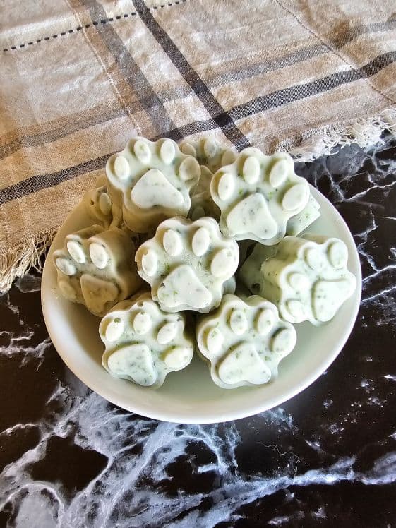 Paw shaped frozen mint dog treats in a white bowl on a dark marble surface next to a linen towel.