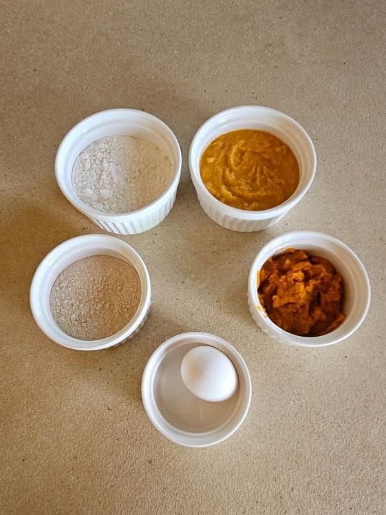 All purpose flour, sweet potato puree, pumpkin puree, an egg, and whole wheat flour in individual white bowls.
