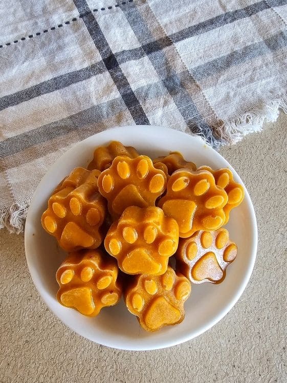 Paw shaped frozen pumpkin banana dog treats in a white bowl next to a linen towel.