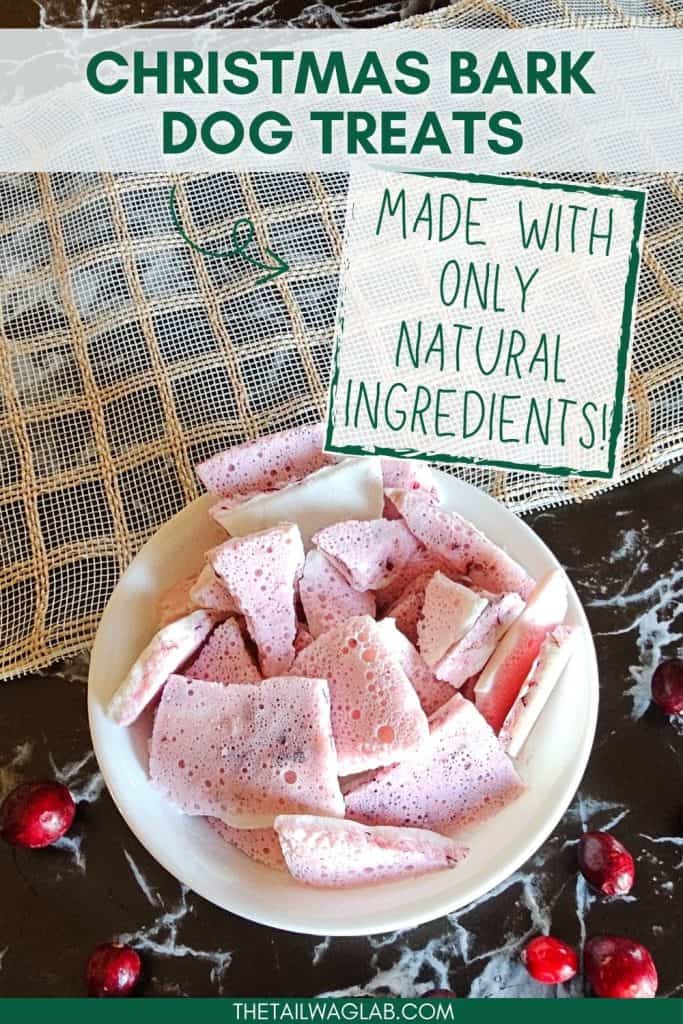 A white bowl filled with pink, foam-like Christmas bark dog treats sits on a marble surface, surrounded by cranberries and featuring a label that states these treats are made with only natural ingredients.