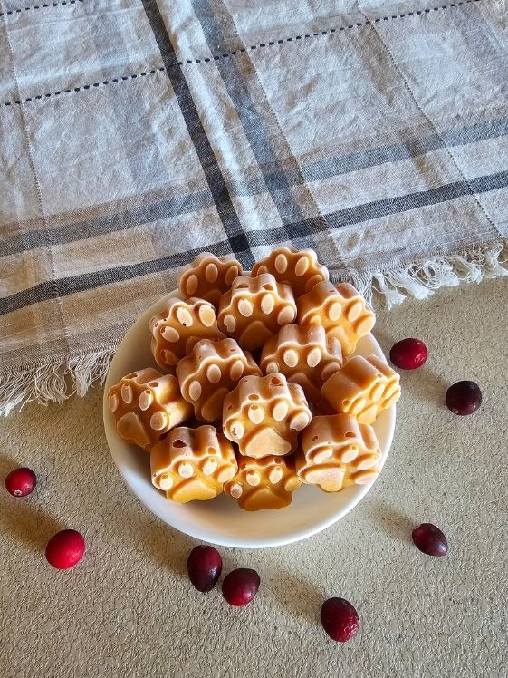 Paw shaped frozen pumpkin cranberry dog treats in a white bowl next to a linen towel with cranberries scattered around the bowl.