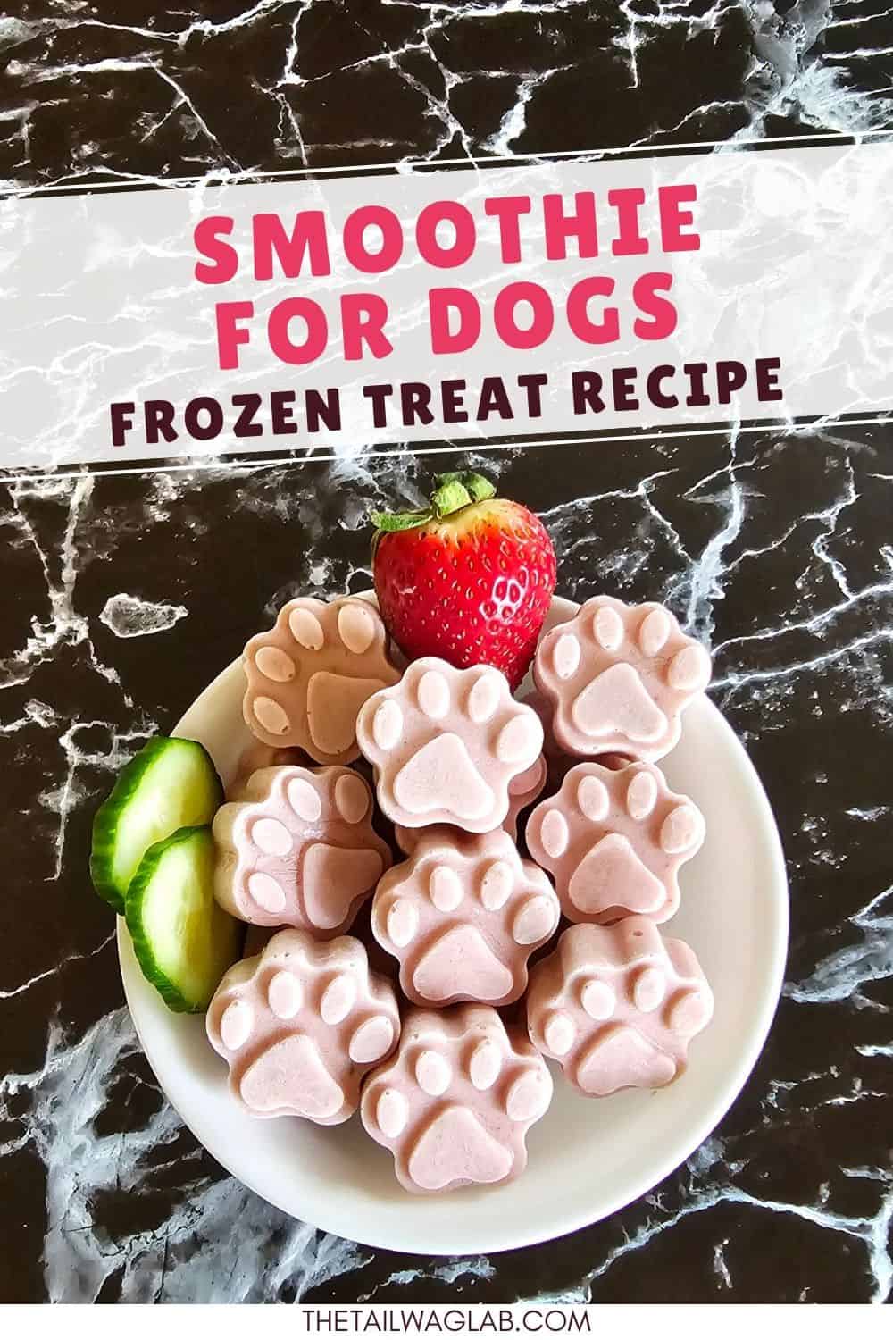 A plate of paw-shaped frozen dog treats made with strawberry yogurt is displayed alongside a fresh strawberry and cucumber slices on a marble surface.