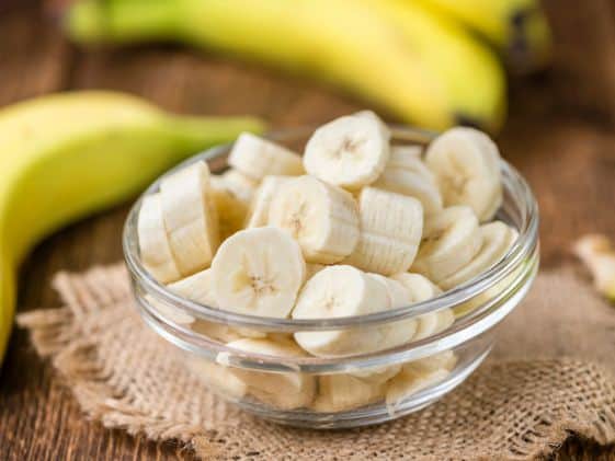A glass bowl of banana pieces next to a few whole bananas.