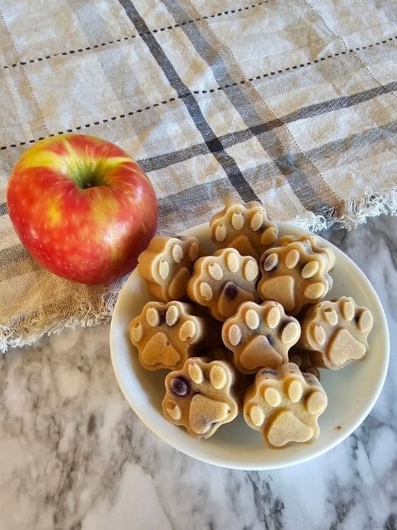 Paw shaped ice cream for dogs in a white bowl next to an apple on a linen towel.