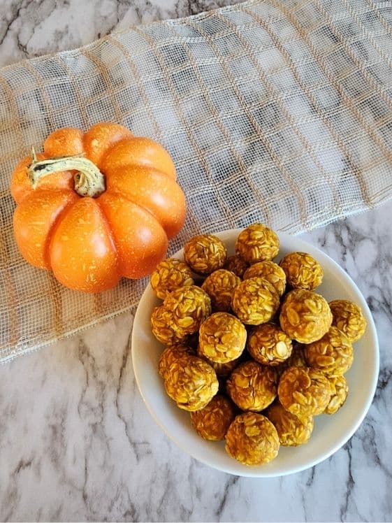 No bake peanut butter pumpkin dog treat balls in a white bowl next to a small pumpkin.