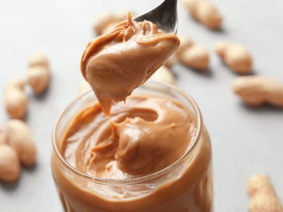 Creamy peanut butter in a glass with a spoon, surrounded by scattered peanut pieces.