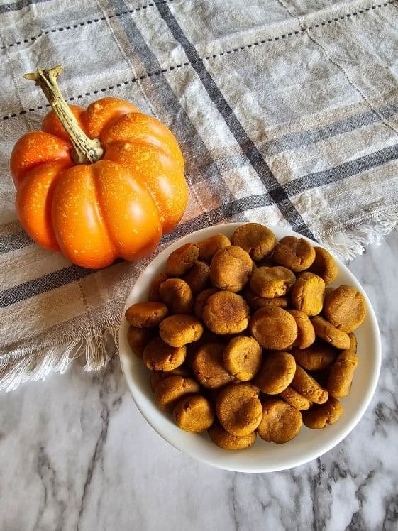 Peanut butter and pumpkin soft baked dog treats in  a white bowl next to a small pumpkin on a linen towel.