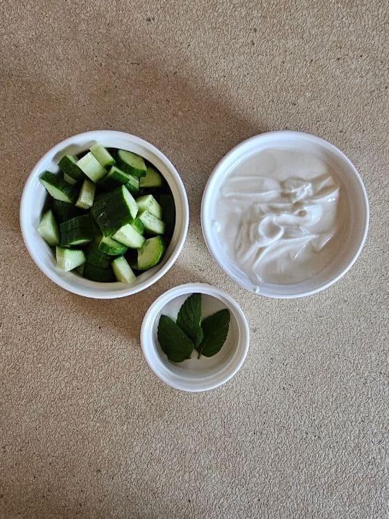 Chopped cucumber, plain Greek yogurt, and mint leaves in individual white bowls.