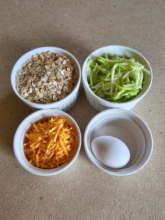 Oats, shredded zucchini, shredded cheddar cheese, and an egg in individual white bowls.