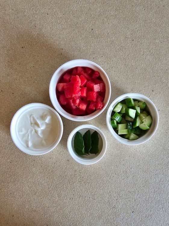 Plain Greek yogurt, chopped watermelon, chopped cucumber, and mint leaves in individual white bowls.