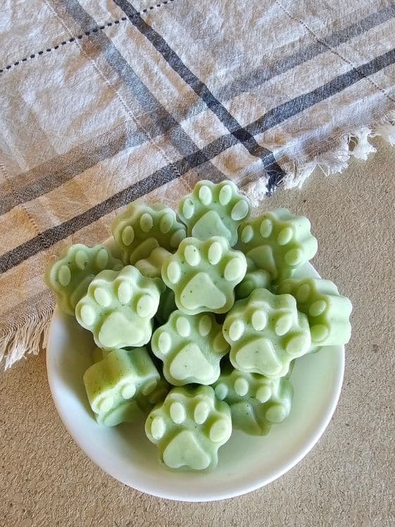 Paw shaped cucumber honeydew frozen dog treats in a white bowl next to a linen towel.