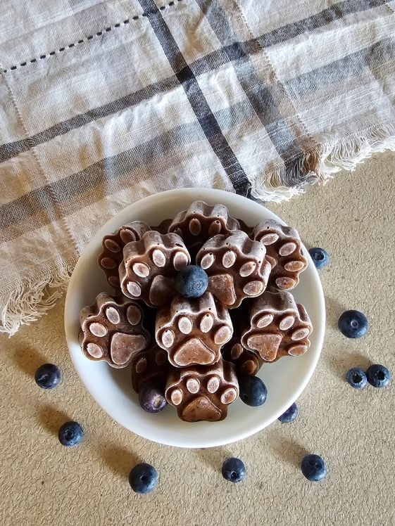 Paw shaped frozen blueberry pumpkin dog treats in a white bowl surrounded by blueberries next to a linen towel.